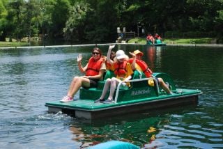 A group of people sitting on a green paddle boat on a lake, waving. 